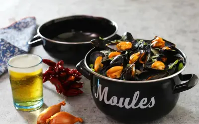 Un bicchiere di birra dorata accanto a cozze fumanti in una pentola nera, con peperoncini secchi e scalogni. Un perfetto abbinamento di birra e pesce.