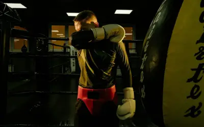 A boxer wipes sweat from his face in a dimly lit boxing ring. He's wearing gloves and a red cup. This image makes you wonder how long do boxers rest after a fight.