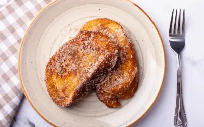 Deux tranches dorées de pain perdu au four, saupoudrées de sucre et de cannelle, sur une assiette avec une serviette à carreaux et une fourchette.