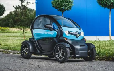 Niebiesko-czarny Renault Twizy, mały samochód elektryczny, zaparkowany na tle niebieskiego budynku i zieleni.