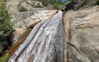 El agua desciende por la roca en la cascada del Hornillo, rodeada de vegetación y rocas graníticas bajo un cielo azul.
