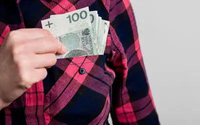 Ręka wkłada banknoty 100 zł do kieszeni koszuli w kratę. Może to symbolizować wynagrodzenie ryczałtowe.