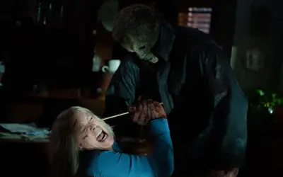 Michael Myers w masce i czarnym płaszczu trzyma nóż nad krzyczącą Laurie Strode. To przerażający halloweenowy finał.