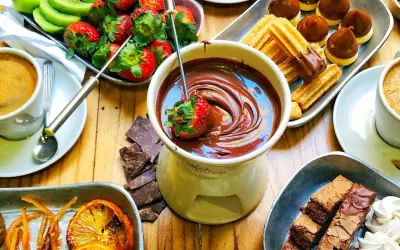 Jak przygotować czekolady do fontanny: truskawka zanurzona w płynnej czekoladzie, obok kawa, ciastka, owoce i brownie.