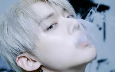 Młody mężczyzna z jasnymi włosami, jeden z członków popularnych k-pop zespołów męskich, wydmuchuje dym.