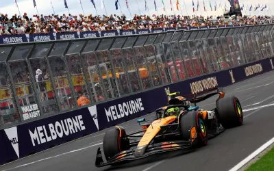 Un coche de Fórmula 1 naranja y negro acelera en Melbourne. Los aficionados vitorean. ¡Qué ganas de que empiece la Fórmula 1 2026!