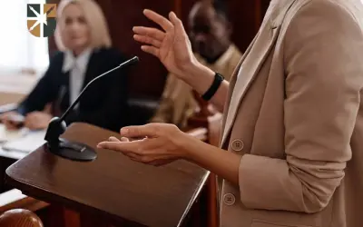 Oskarżyciel posiłkowy przemawia przy mównicy, gestykulując. W tle siedzą sędziowie.