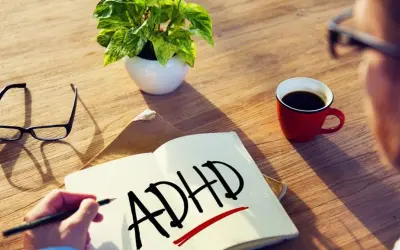 Osoba zmagająca się ze spektrum ADHD pisze w zeszycie. Obok leżą okulary, filiżanka kawy i roślina.