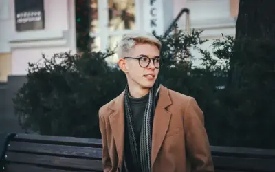 Młody mężczyzna w okularach i płaszczu prezentuje sportową elegancję, siedząc na ławce wśród zieleni.
