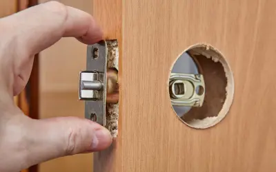 Dłoń wsuwa metalowy mechanizm zamka do wyciętego otworu w drewnianych drzwiach.