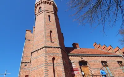 Ceglana latarnia morska w Ustce z charakterystyczną wieżą i budynkiem. Na balkonie stoi kobieta.