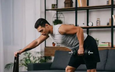 Mężczyzna wykonuje trening siłowy w domu, podnosząc hantle, opierając się o krzesło.