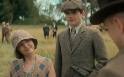 Giovane donna con cappello a scacchi e gilet a rombi, accanto a un uomo con berretto e giacca di tweed. Sullo sfondo, i sette quadranti di un campo.