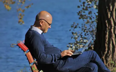 Mężczyzna w okularach siedzi na ławce nad jeziorem, pracując na laptopie. Planuje swoje ubezpieczenie emerytalne, ciesząc się spokojem natury.