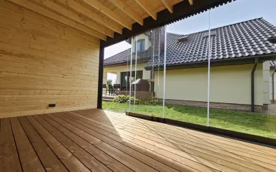 Drewniana altana przy ścianie domu z przeszklonymi panelami, wychodząca na zielony ogród i dom z brązowym dachem.