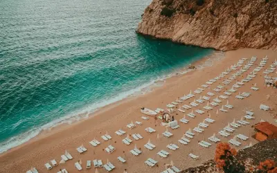 Plaża w Turcji, turkusowe morze, piaszczysty brzeg z rzędami leżaków i parasoli, skaliste wybrzeże.
