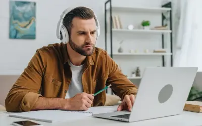 Mann mit Kopfhörern lernt online, nutzt Laptop und Notizbuch. Er wirkt konzentriert, während er mit einem Stift tippt. Ein typisches Bild für das heutige "Home-Office"-Zeitalter, das viele Anglizismen mit sich bringt.