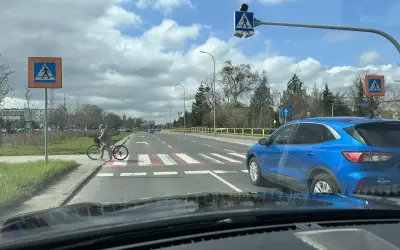 Niebezpieczne wyprzedzanie na pasach na drodze dwupasmowej. Niebieski SUV omija rowerzystkę stojącą przy przejściu.