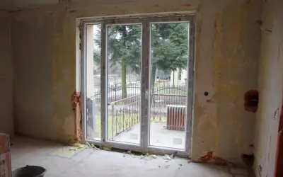 Niedokończona ściana z oknem balkonowym, widok na drzewa i ogrodzenie.