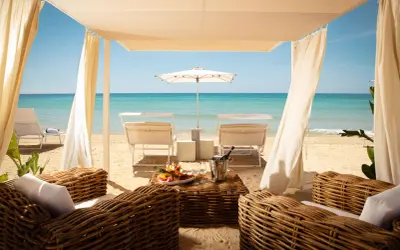 Lussuosa area lounge con divani in vimini, tavolino con frutta e champagne, lettini e ombrellone su una spiaggia di sabbia dorata. Un'oasi tra i migliori beach clubs Puglia.