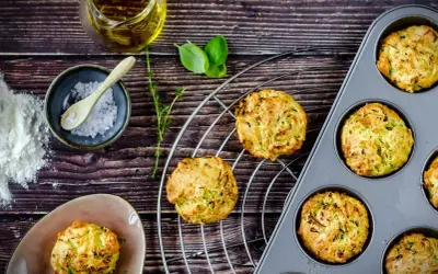Goldbraune Zucchini-Muffins auf einem Gitter, einige in einer Muffinform. Zutaten wie Öl, Salz und Kräuter liegen bereit.