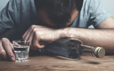 Mężczyzna z głową opartą o butelkę, obok kieliszek z alkoholem. Wygląda na to, że doświadcza zatrucia alkoholowego.