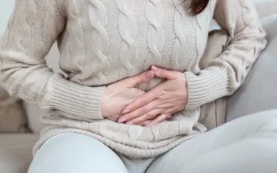 Kobieta w beżowym swetrze trzyma się za brzuch, odczuwając bolesne rozpychanie dziecka w brzuchu.