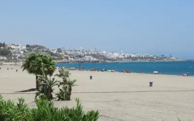 La **playa de la Mena** se extiende con arena dorada y palmeras. Al fondo, un pueblo blanco se asoma al mar azul.