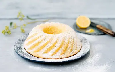 En vackert pudrad sockerkaka med citron, serverad på en mörk tallrik med en citronskiva och en kniv i bakgrunden.