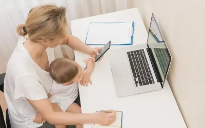 Kobieta z dzieckiem na kolanach sprawdza w telefonie, ile płatny urlop macierzyński. Obok laptop i notatnik.