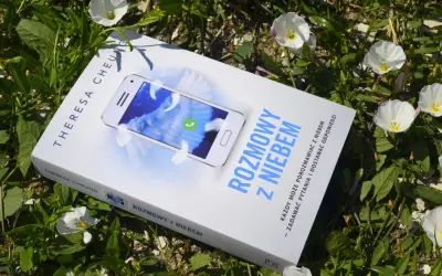 Książka "Rozmowy z niebem" leży wśród zieleni i białych kwiatów. Na okładce telefon z napisem "Niebo" i piórami.