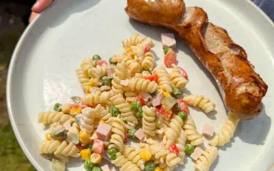 Gegrillte Wurst mit einem cremigen Nudelsalat als Beilage, garniert mit Erbsen, Mais und gewürfeltem Schinken.