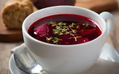 Aromaticzny barszcz czerwony domowy, z kawałkami buraka i ziołami, podany w białej filiżance z łyżeczką.
