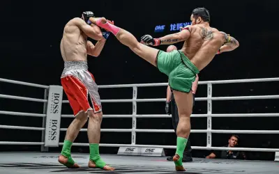 A Muay Thai fighter in green shorts lands a powerful kick to his opponent's head. The opponent, in red shorts, braces for impact, his foot nearly touching the canvas.