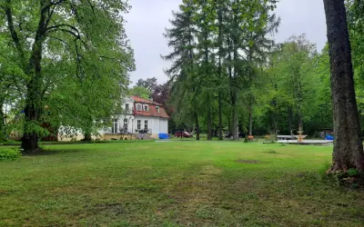 Biały dworek z czerwonym dachem otoczony zielenią. W oddali widać fontannę i drzewa. To urokliwa **gierłoż**.