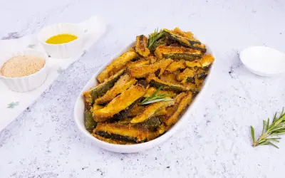 Zucchine gratinate al forno, dorate e croccanti, servite in un piatto ovale con rosmarino fresco. Accanto, ciotoline con pangrattato e olio.