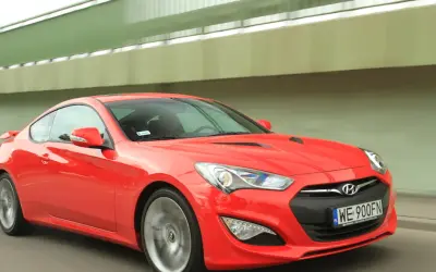 Czerwony Hyundai Genesis Coupe 3.8 V6 w ruchu, z widocznym kierowcą i tablicą rejestracyjną WE 900FN.
