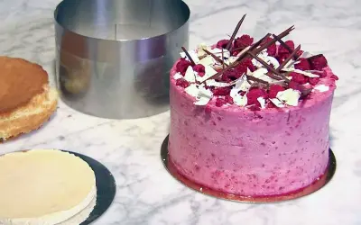 En rosa hallontårta med choklad och vita dekorationer. Bredvid ligger en bakring och delar av en tårta, redo för ett nytt tårta recept.
