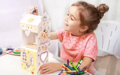 Uśmiechnięta dziewczynka ozdabia kartonowy domek, który jest świetną zabawka dla sześciolatków. Kolorowe kredki i farby czekają na użycie.