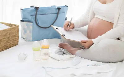 Gravid kvinna skriver på en lista för packlista bb. Bredvid ligger en blå väska, korg med kläder, nappflaskor och bebiskläder.