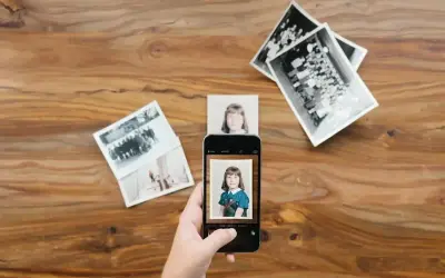 Skan ze zdjęcia: dłoń trzyma smartfon, który fotografuje zdjęcie dziewczynki. Wokół leżą stare fotografie.