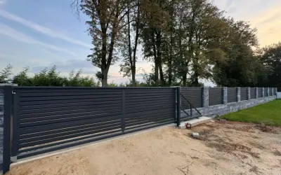 Nowoczesne, ciemnoszare ogrodzenie z przesuwną bramą. Widać kamienny murek i drzewa w tle. Odległość ogrodzenia od drogi gminnej wewnętrznej jest niewielka.