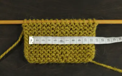 Knitted swatch with a measuring tape across it, showing stitches on wooden needles.