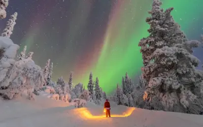 Un uomo con lanterna, vestito da Babbo Natale, ammira l'aurora boreale in Lapponia. Un paesaggio innevato e magico, perfetto per chi cerca dove andare in Lapponia.