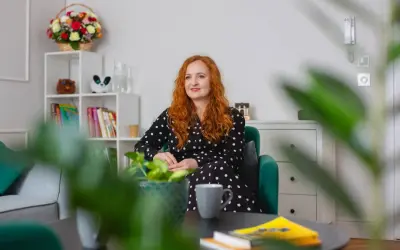Kobieta z rudymi włosami w czarnej sukience w kropki siedzi w zielonym fotelu. Na stoliku leżą książki, a w tle widać regał z dekoracjami. To idealne miejsce na rozmowy z conners 3.
