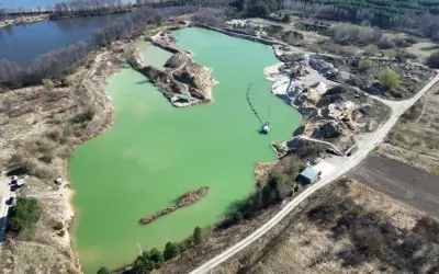 Widok z lotu ptaka na turkusowe jezioro zajączkowskie, otoczone piaszczystymi skarpami i zielenią. Widać sprzęt wydobywczy i łódź.