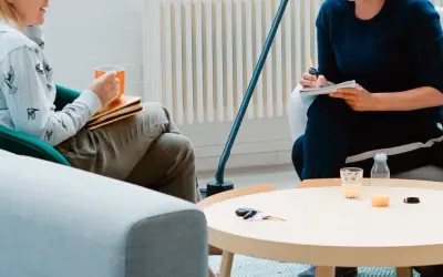 Dwie osoby siedzą w przytulnym pokoju, jedna pije z kubka, druga pisze w notatniku.