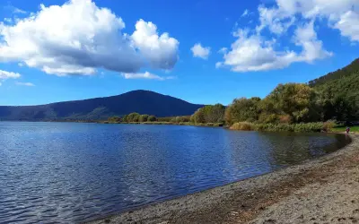 Spiaggia ghiaiosa e acque calme del lago di Vico, con montagne verdi e cielo azzurro punteggiato di nuvole bianche.