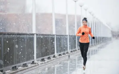 Donna corre sotto la pioggia su un ponte, con fiocchi di neve che cadono. Indossa abbigliamento sportivo arancione e nero.