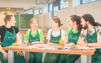 Fünf junge Menschen in grünen Schürzen lachen und arbeiten an einem Holztisch. Hier beginnt ihre kostenfreie Ausbildung Ergotherapie.